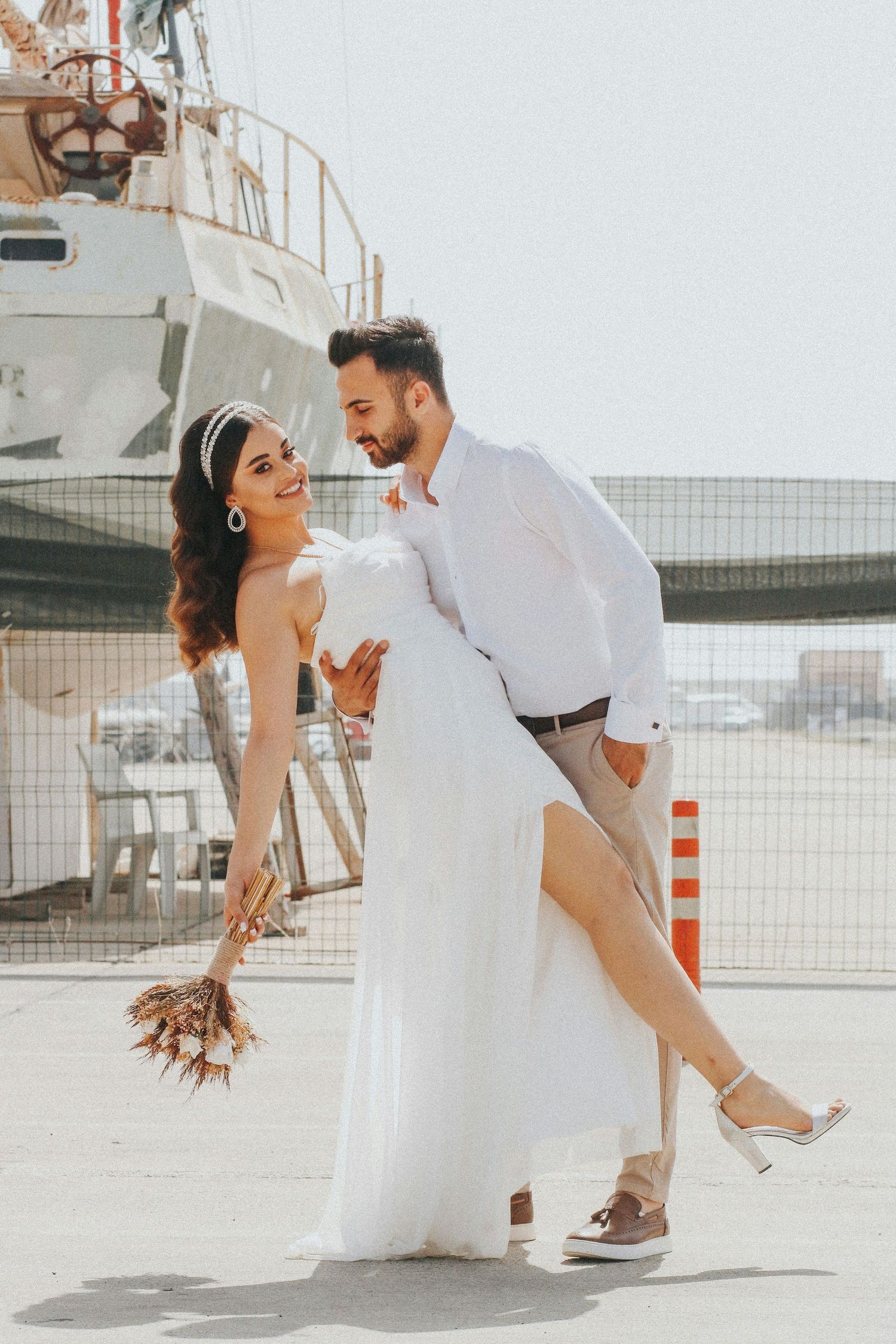 Smiling bride in white dress and groom in white shirt pose playfully near a boat at a sunny harbor.