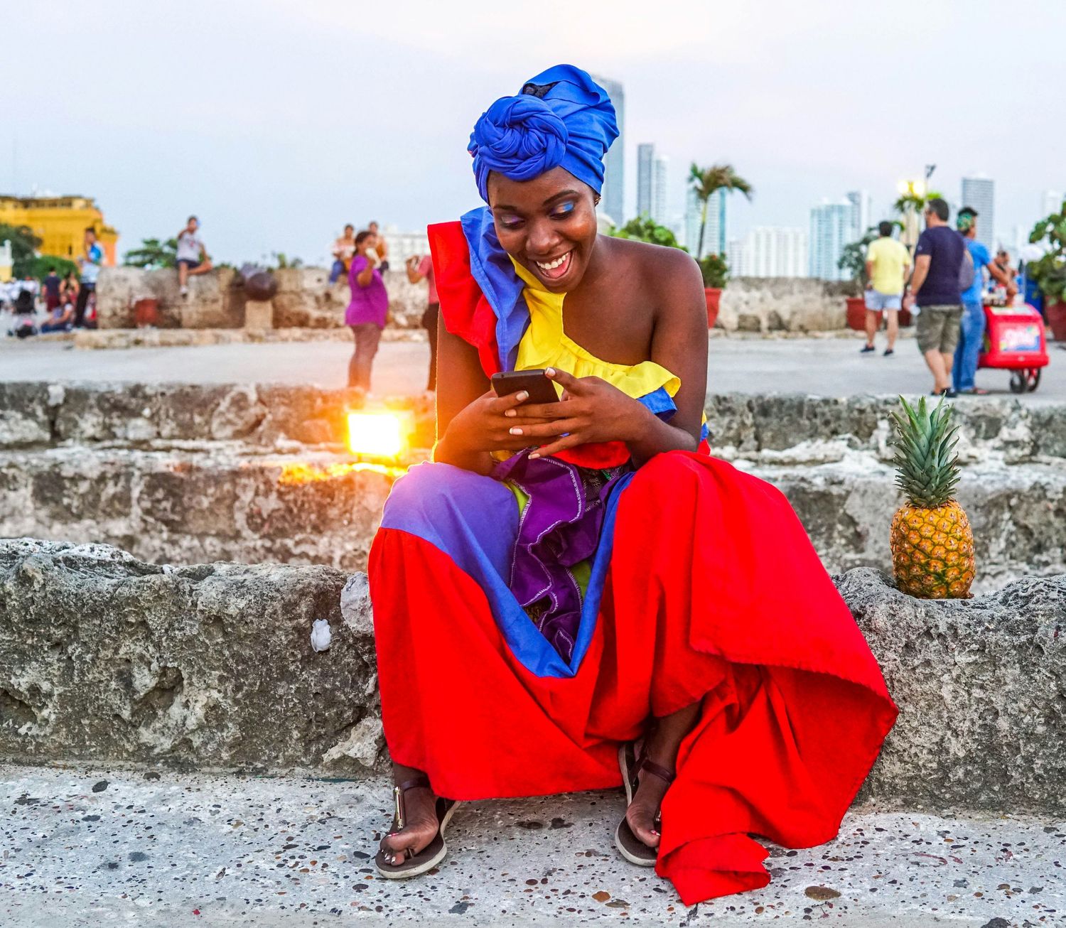 Smiling woman in colorful red and blue dress using smartphone while sitting on stone wall.
