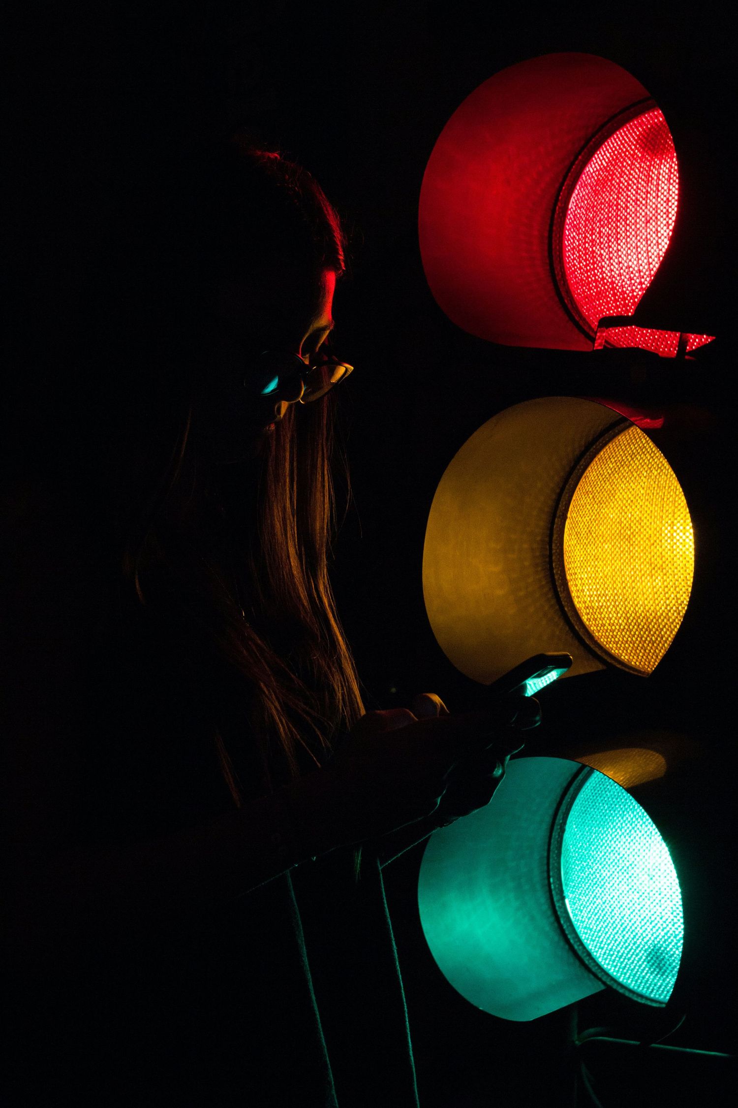 Silhouette of woman standing beside illuminated red yellow and green traffic lights in darkness.