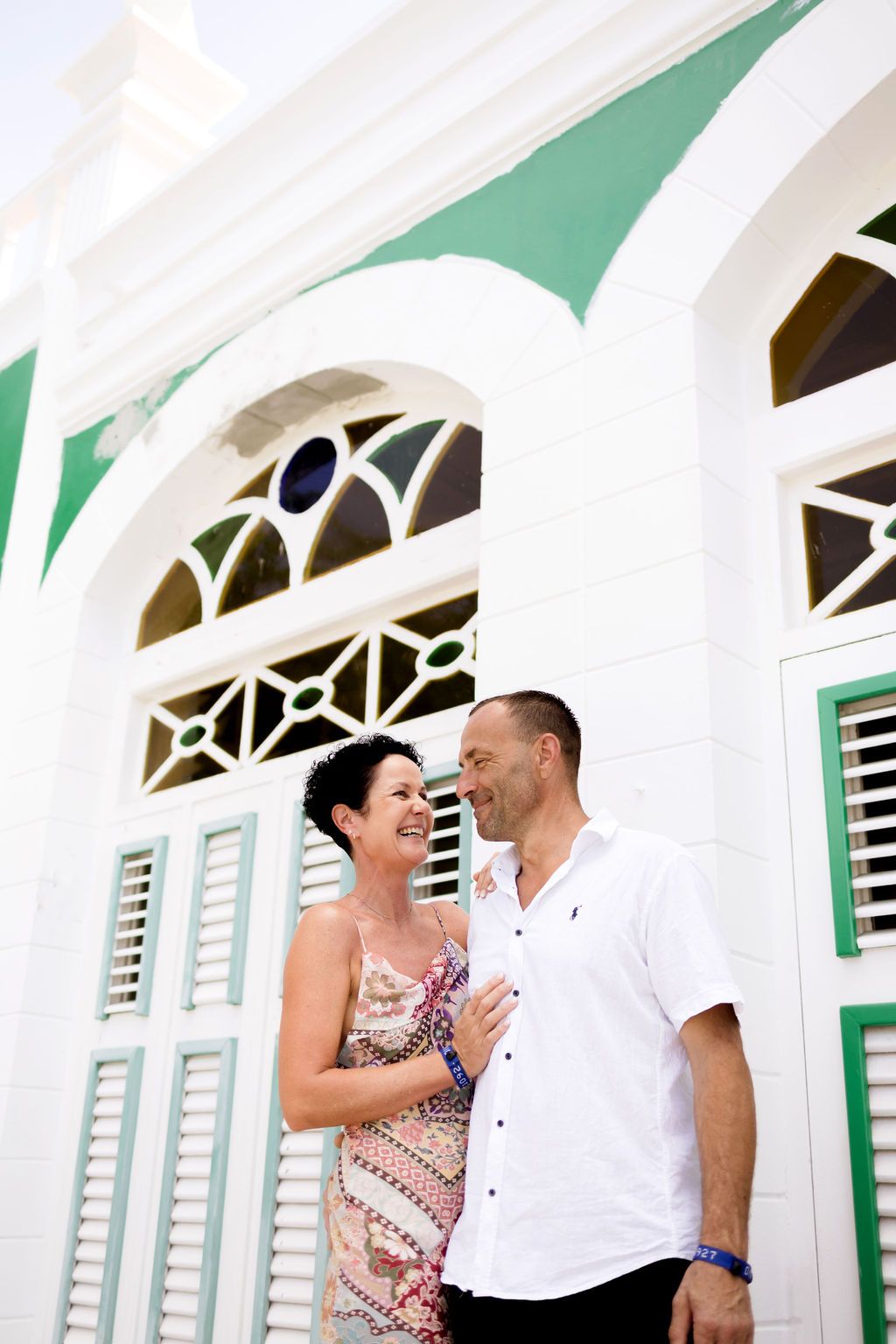 Happy couple laughs together outside a charming white and green colonial-style building in the tropics.