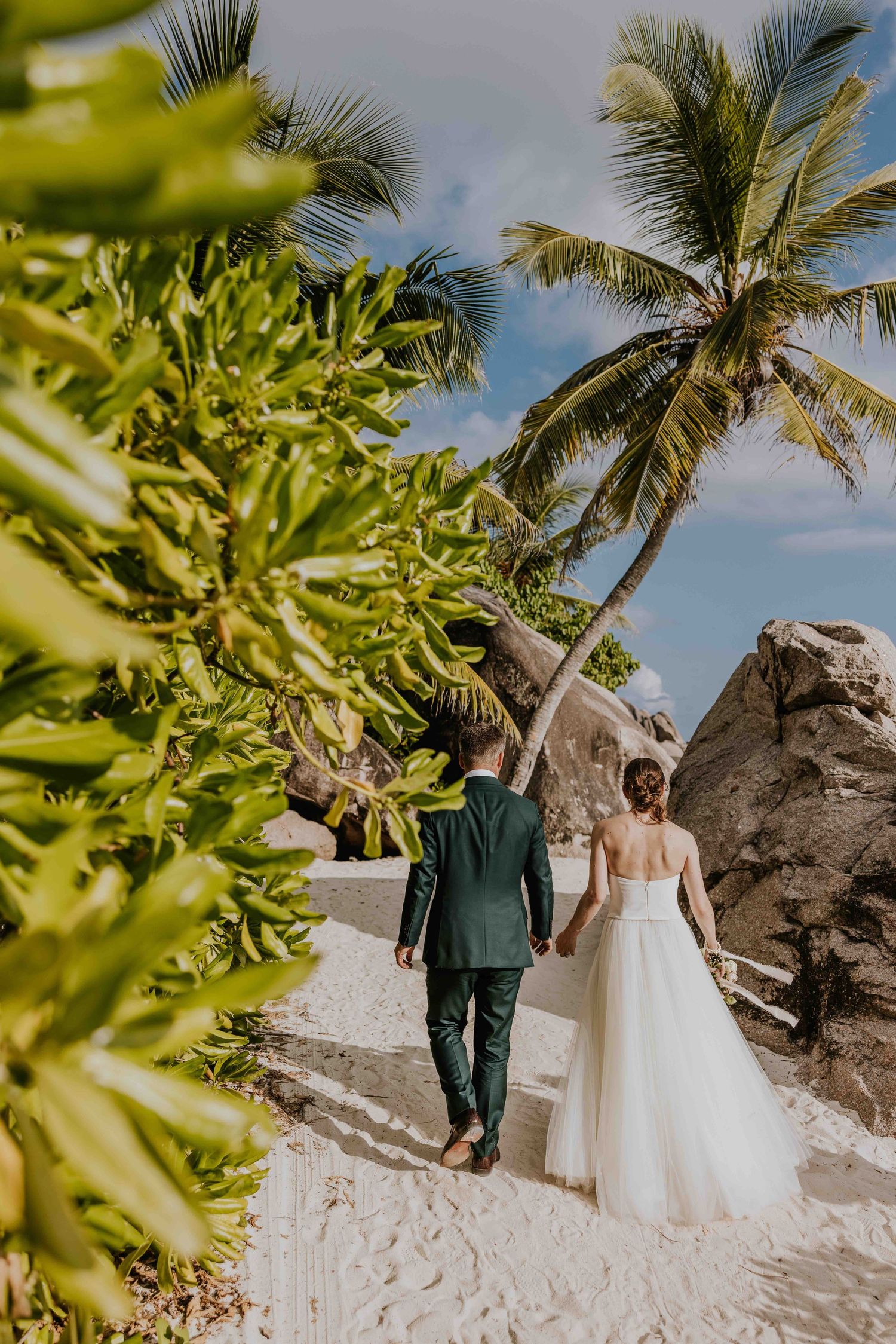 Bride and groom walk hand-in-hand on a tropical white sand beach with palm trees and large rocks.