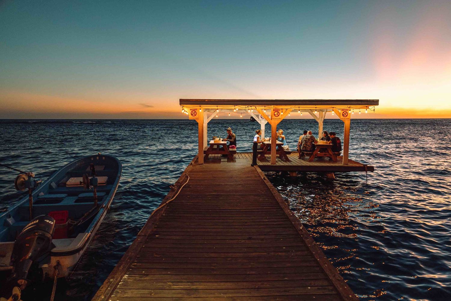 Wooden pier extending over ocean at sunset with illuminated gazebo and boats, creating a serene tropical waterfront scene.