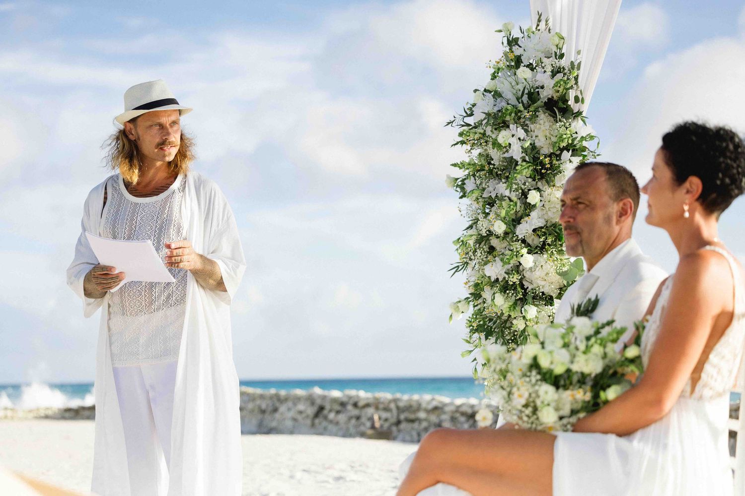 Beach wedding ceremony with white floral arch, officiant reading vows by the ocean.
