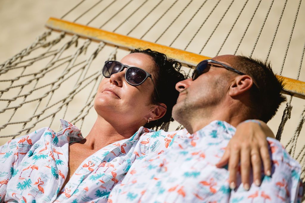 Couple relaxing together in a hammock on a sunny beach, wearing matching floral shirts and sunglasses.