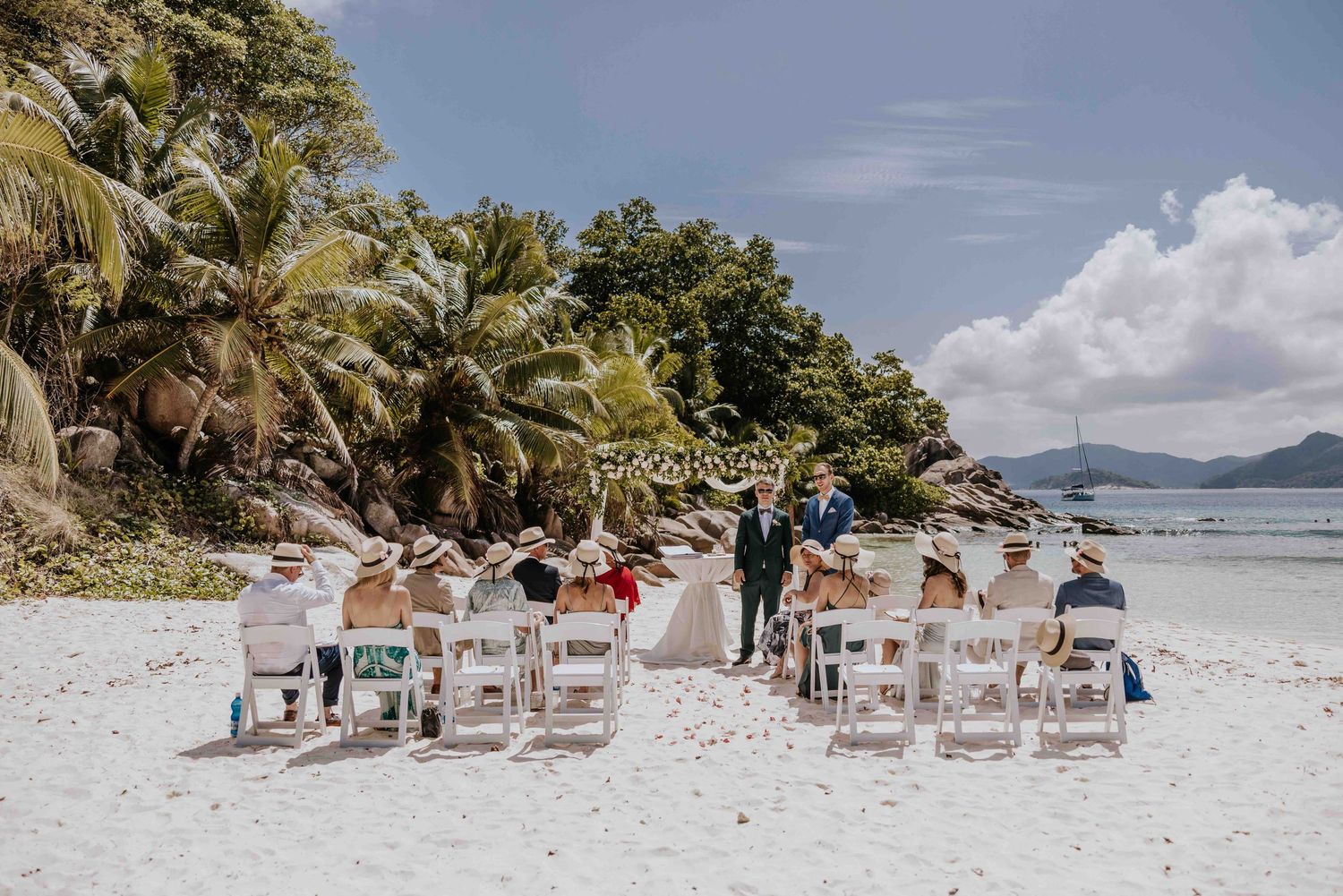 Intimate beach wedding ceremony with small guest gathering on white sand surrounded by tropical palm trees.