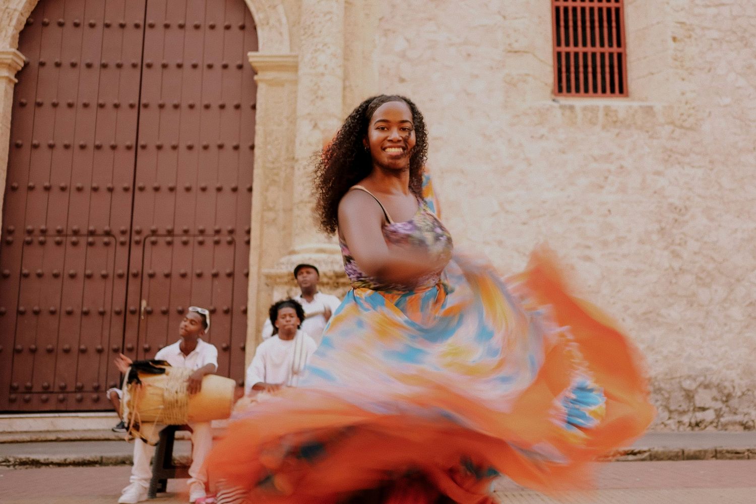 Joyful woman dancing in a colorful swirling dress outside a historic stone building with musicians behind her.