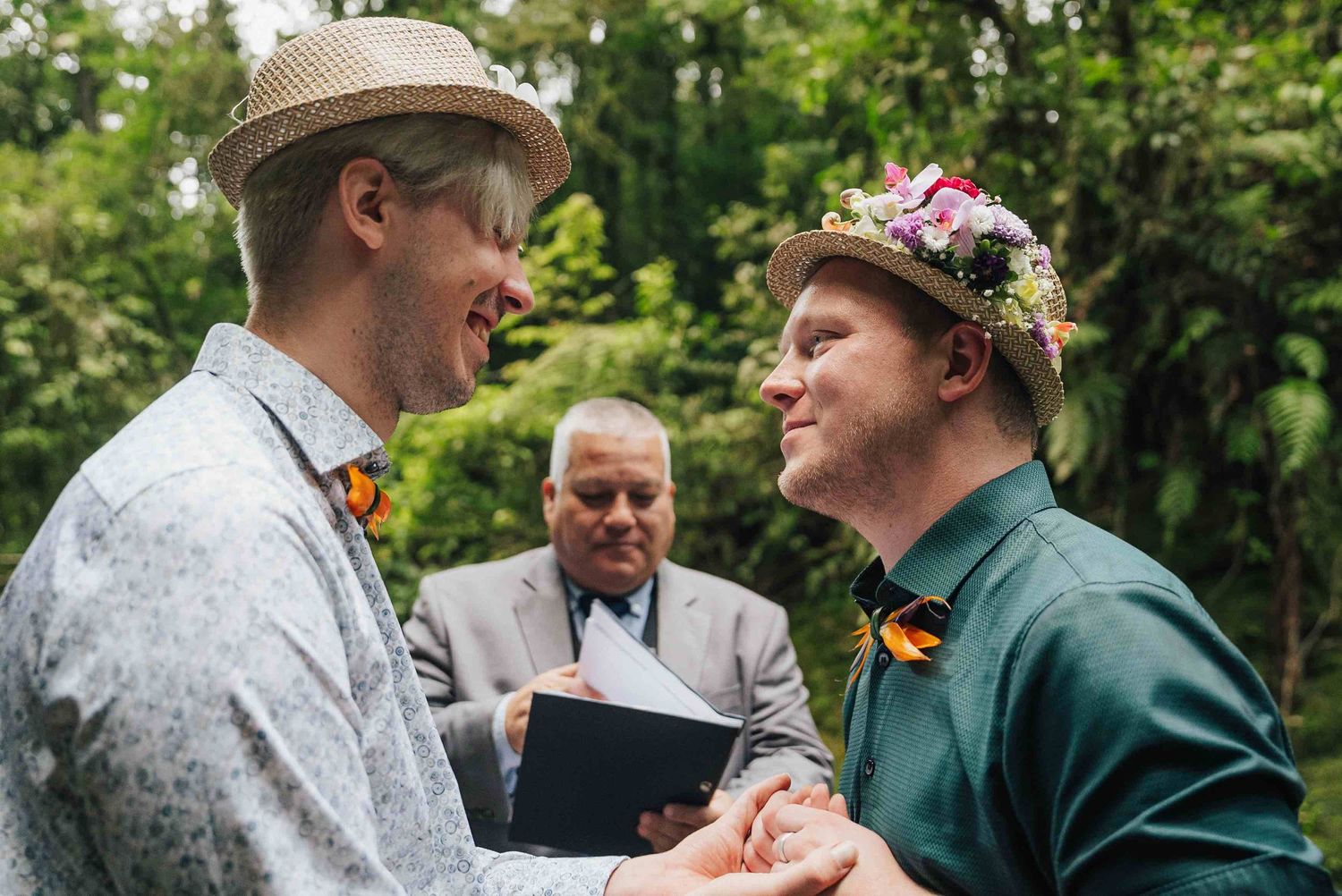 Two grooms exchange vows outdoors, wearing floral hats and bow ties, with an officiant holding a book behind them.