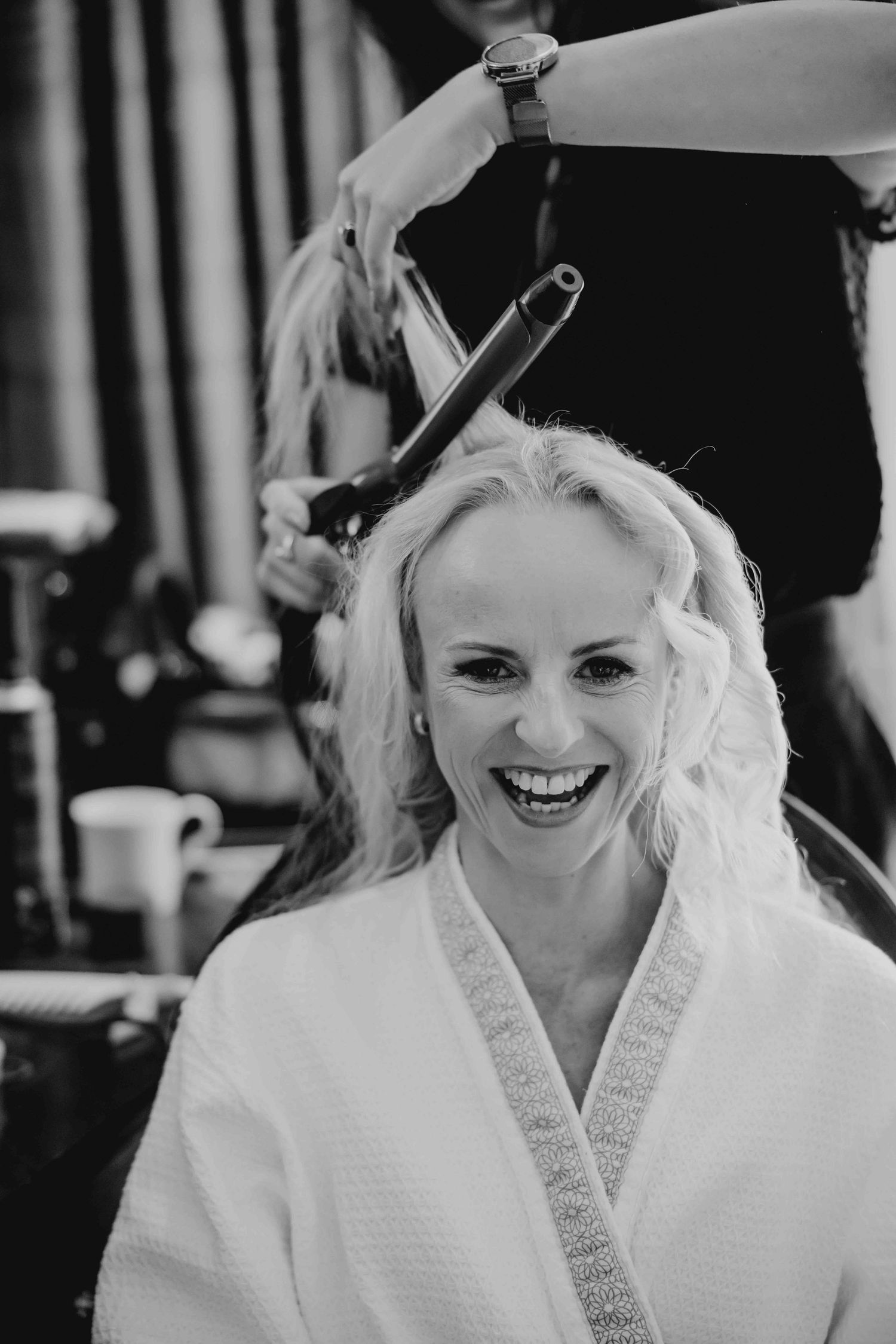 Smiling blonde bride in white robe getting hair styled with curling iron in black and white photo.