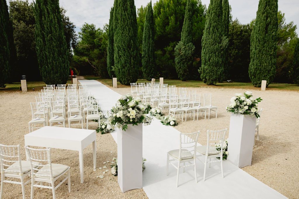 Elegant outdoor wedding ceremony setup with white chairs, floral arrangements, and cypress trees backdrop.