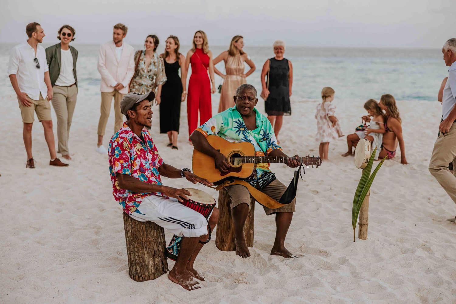 Musicians playing guitar at a beach gathering with guests watching at sunset.