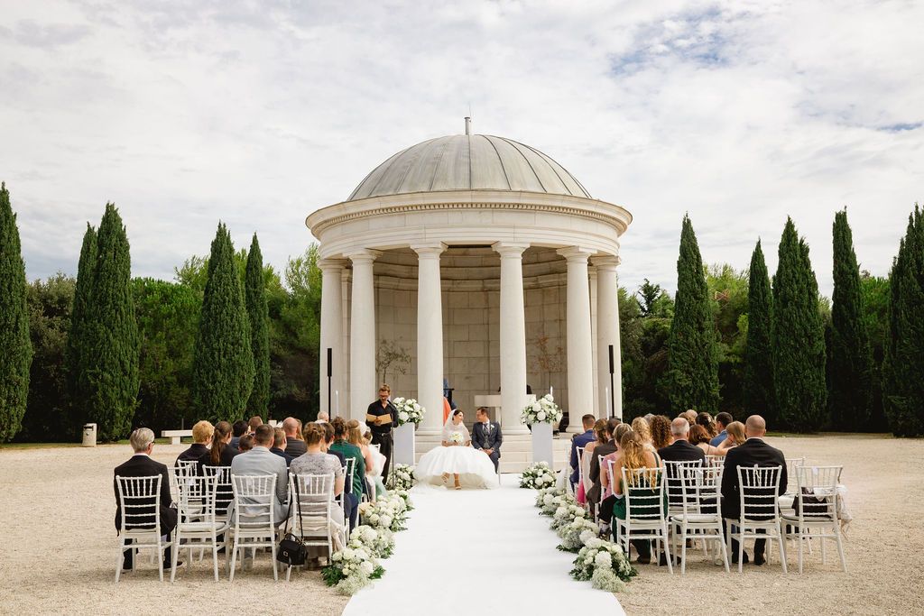 Wedding ceremony at classical rotunda pavilion with white aisle, floral decor, and cypress tree garden.
