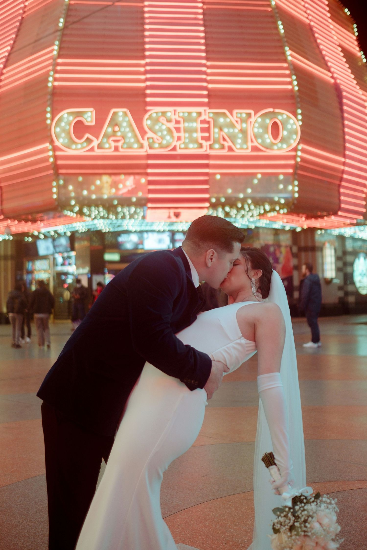 Newlyweds share a passionate kiss in front of a glowing neon Casino sign in Las Vegas at night.