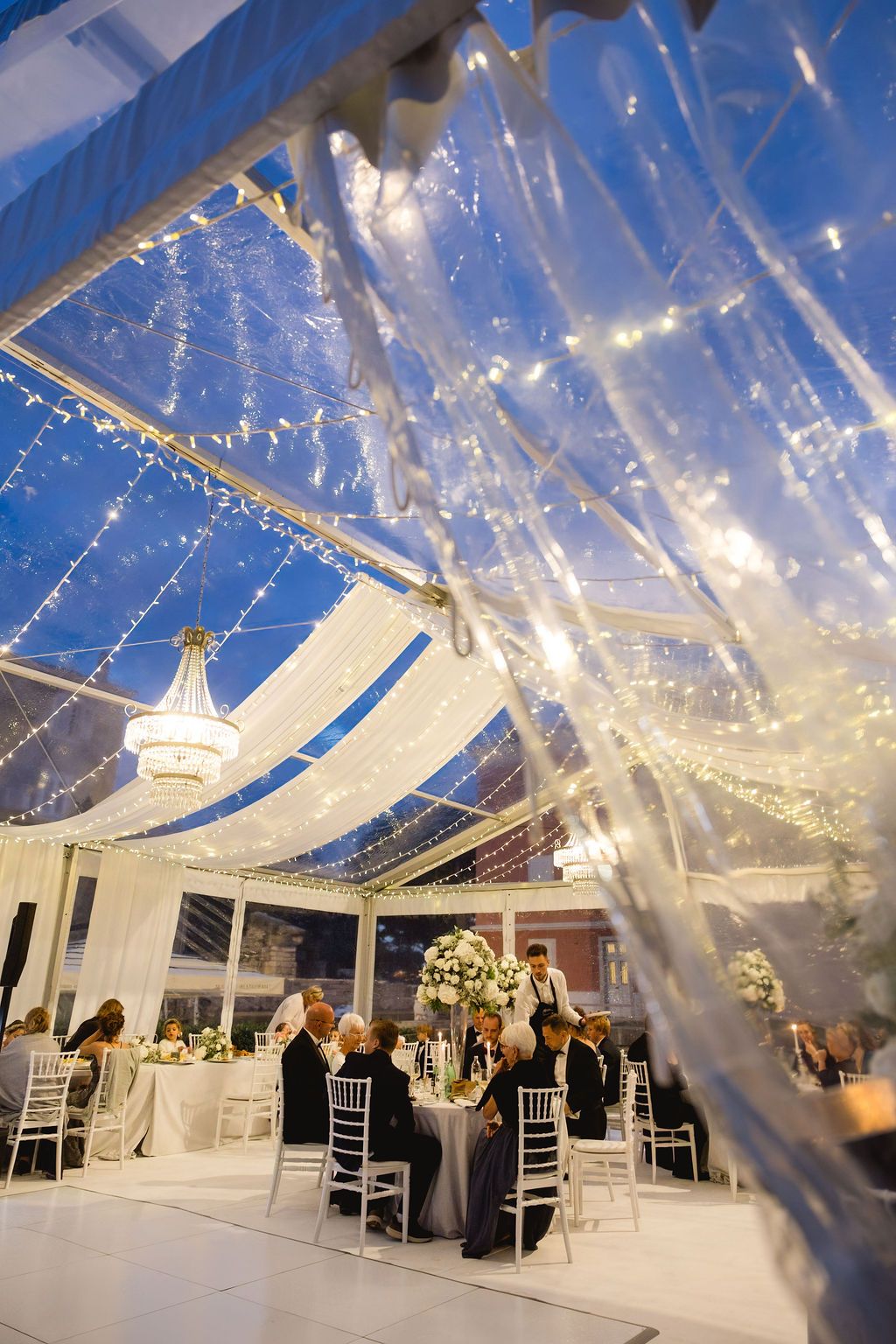 Elegant wedding reception under a clear tent with fairy lights and chandelier against a twilight sky.