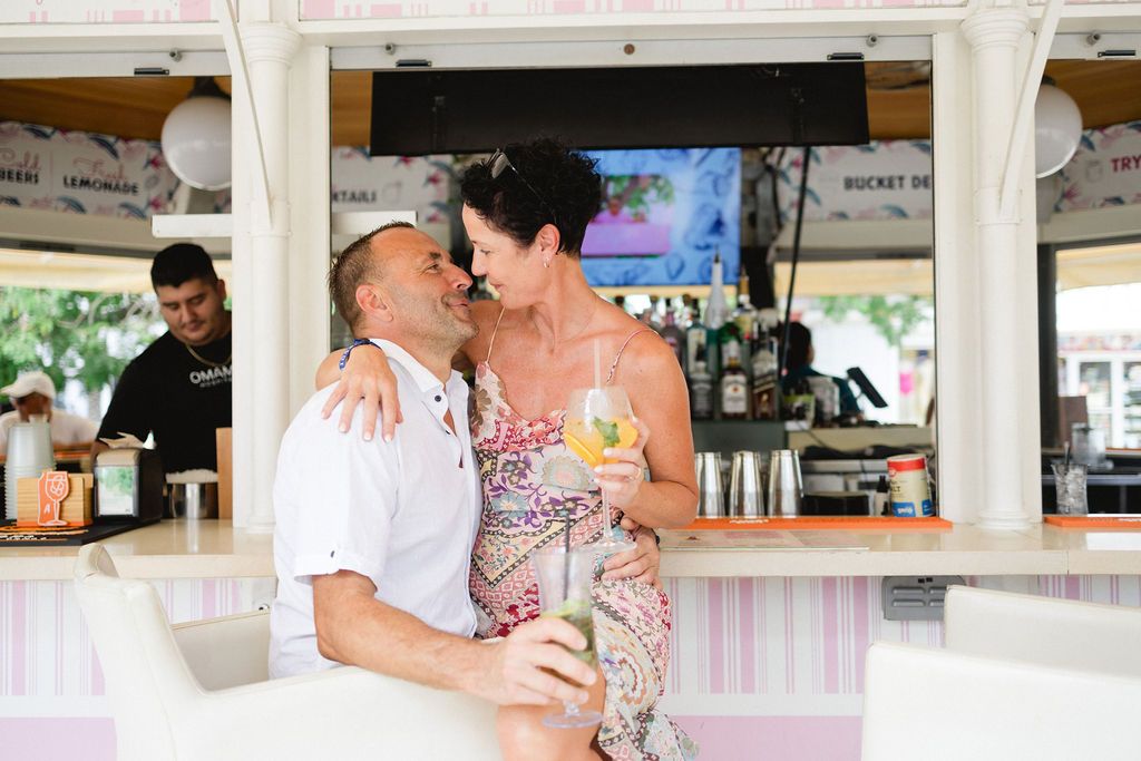 Couple sharing a romantic kiss at an outdoor beach bar while holding a tropical cocktail drink.