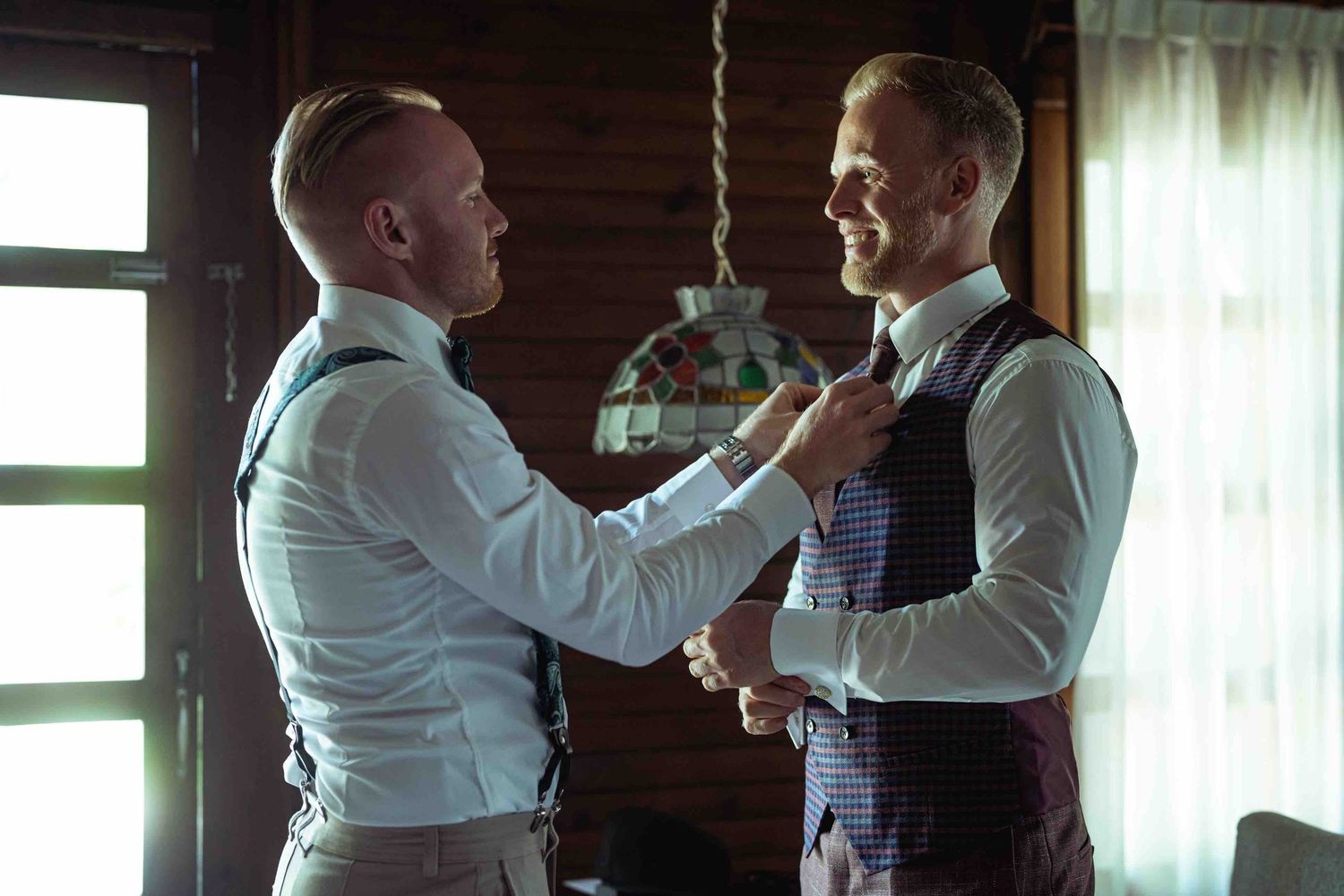 Two men in formal wedding attire adjusting ties and suspenders indoors before a ceremony.