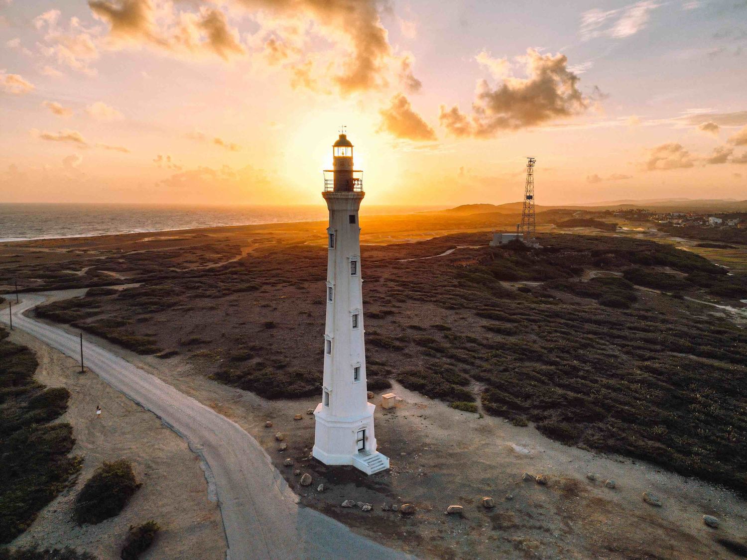 Aerial drone view of a white lighthouse at golden sunset with dramatic orange sky and coastal landscape in Aruba.