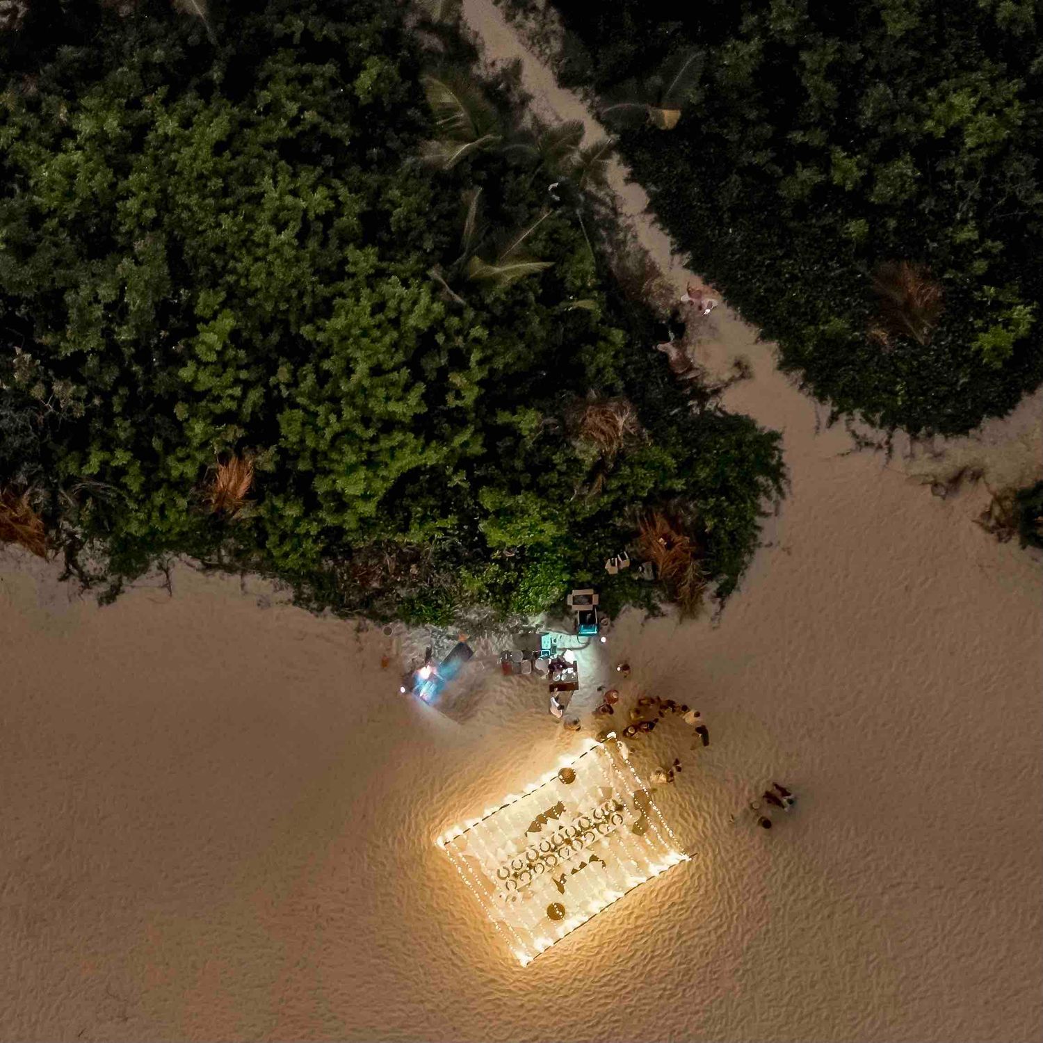 Aerial night view of illuminated beach dining setup near tropical jungle shoreline.