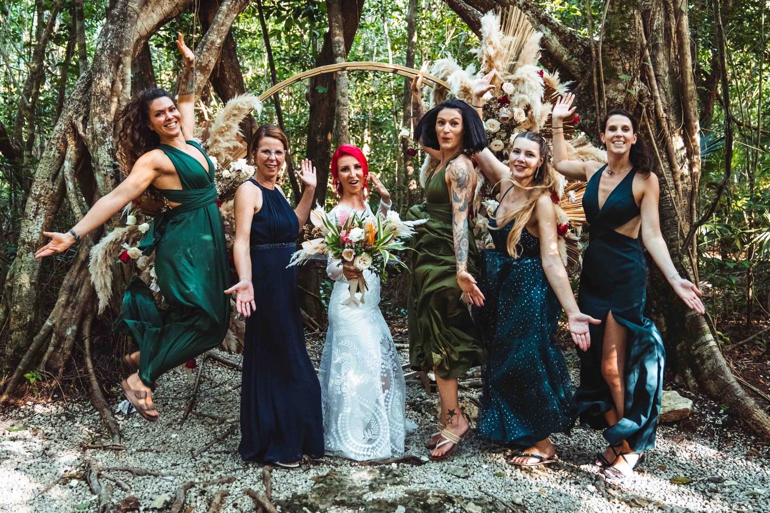 Bride and bridesmaids in green and navy dresses jumping joyfully in a tropical jungle setting.