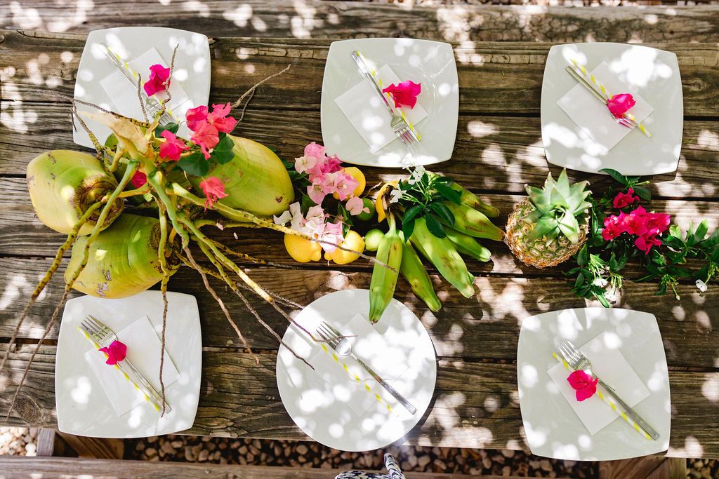 Aerial view of a rustic wooden table set with white plates, pink flowers, and tropical greenery.