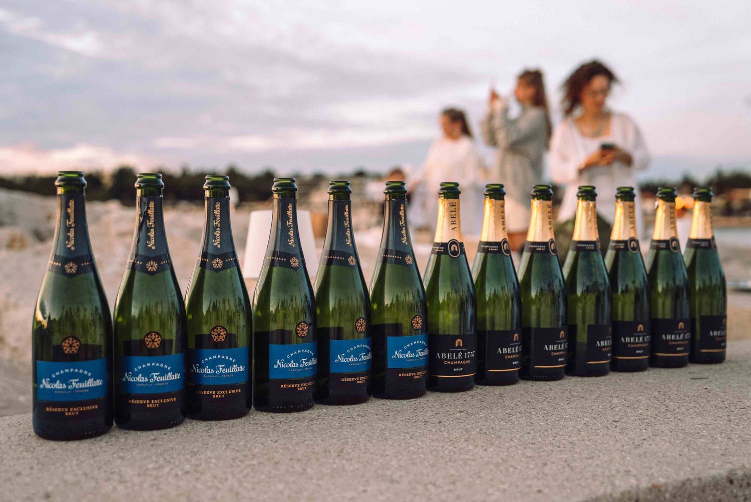 A row of Nicolas Feuillatte champagne bottles lined up on a stone ledge at sunset with people in the background.