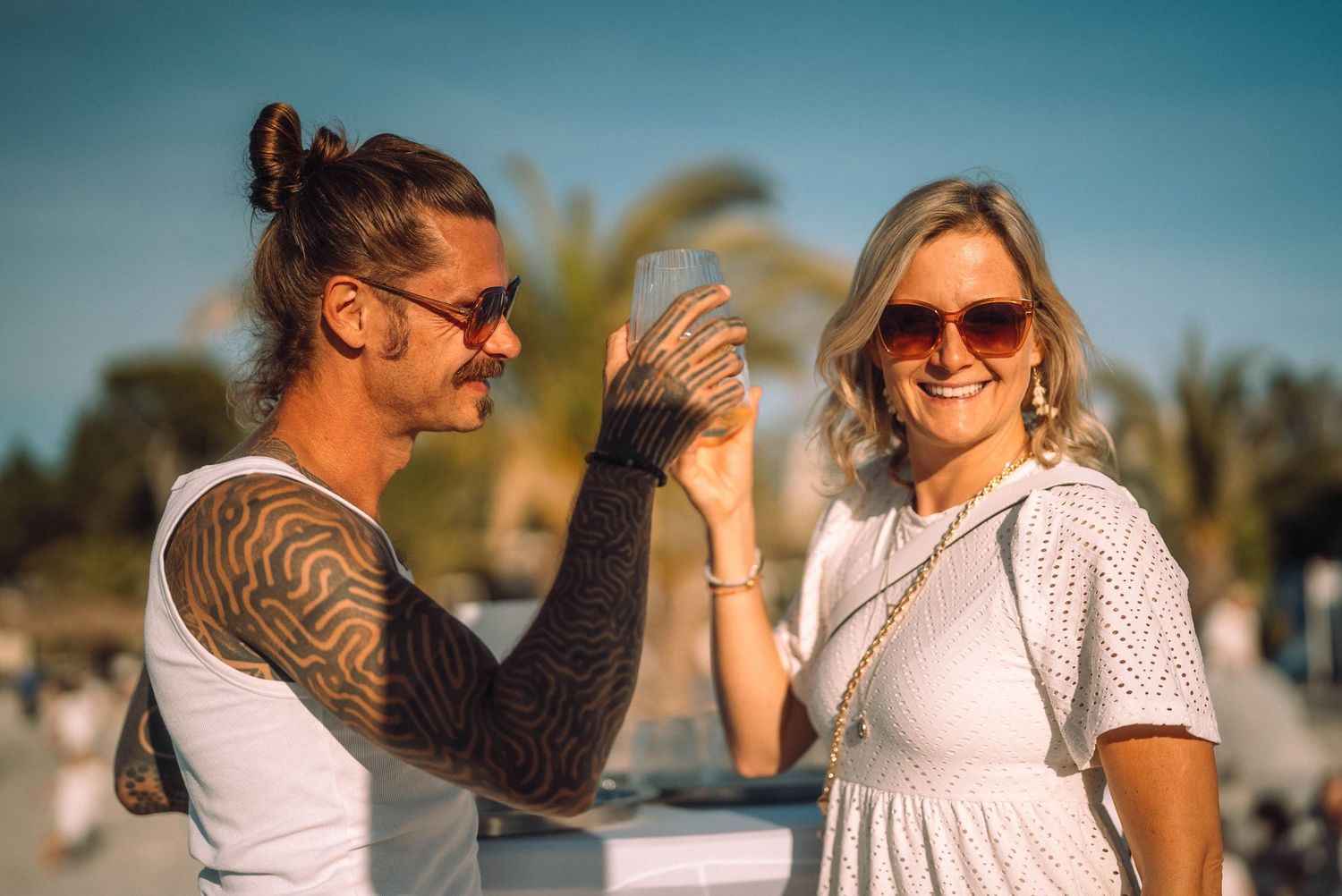 Tattooed man and smiling blonde woman wearing sunglasses enjoying a sunny day outdoors near palm trees.