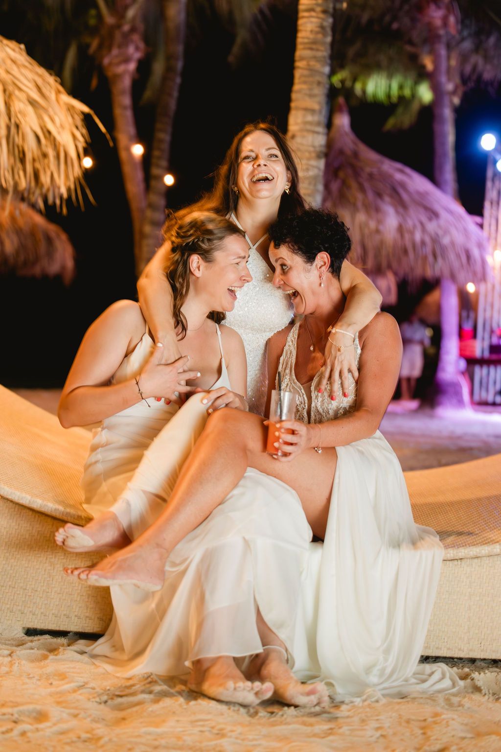 Three joyful women in white dresses laugh and embrace together on sandy beach at night.