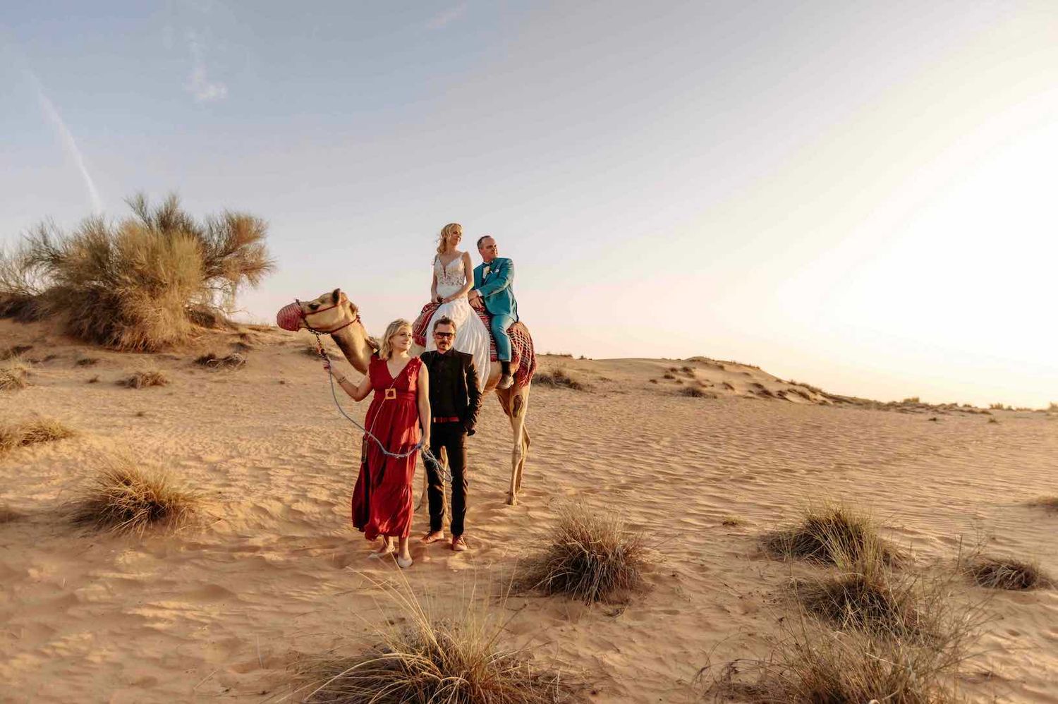 Couple poses with camel riders on golden sand dunes at sunset in a desert landscape.