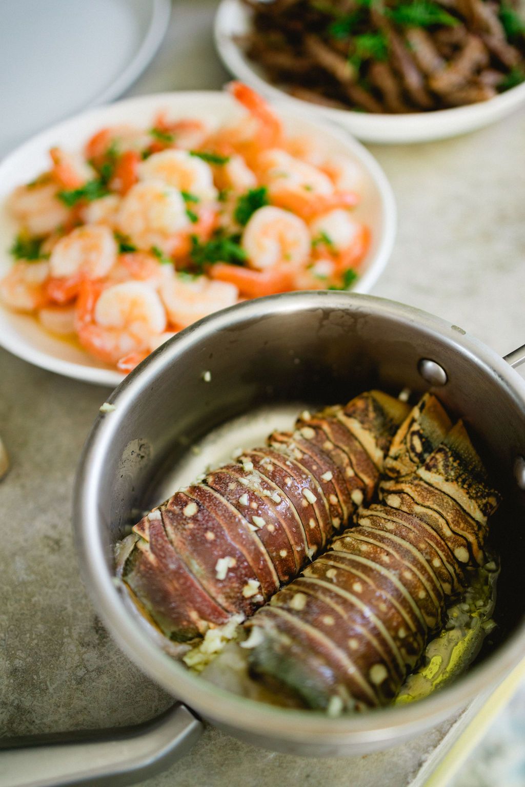 Lobster tail cooking in a pan with shrimp and beef dishes in the background.
