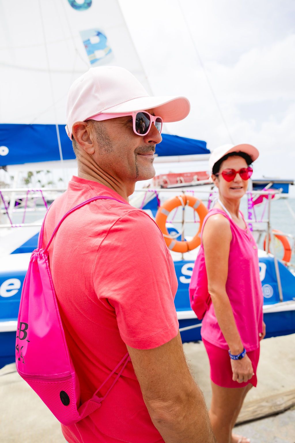 Two people in matching pink outfits and sunglasses stand near a blue sailboat at a sunny marina.