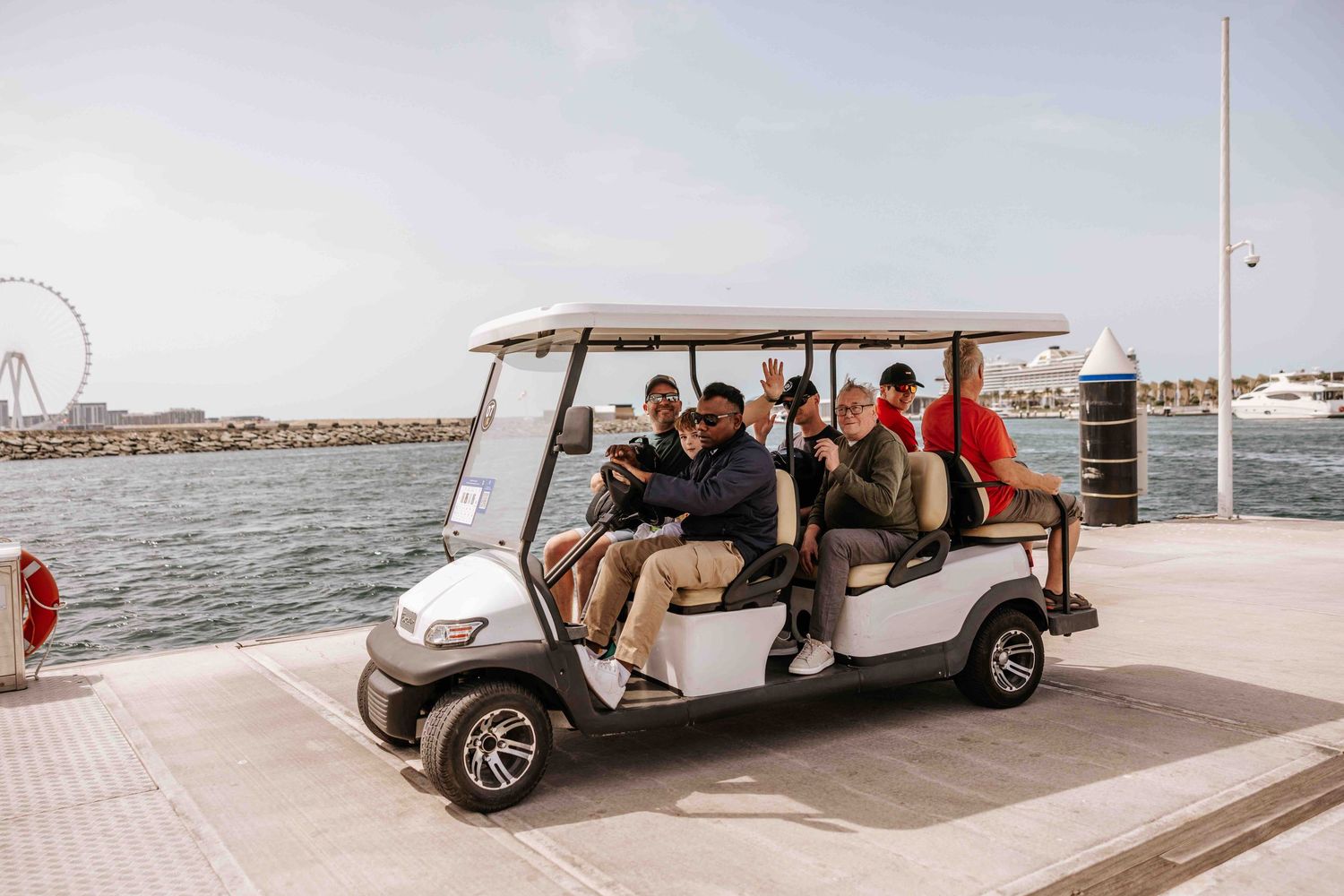 Group of people riding a white golf cart along a waterfront promenade with a Ferris wheel in the background.