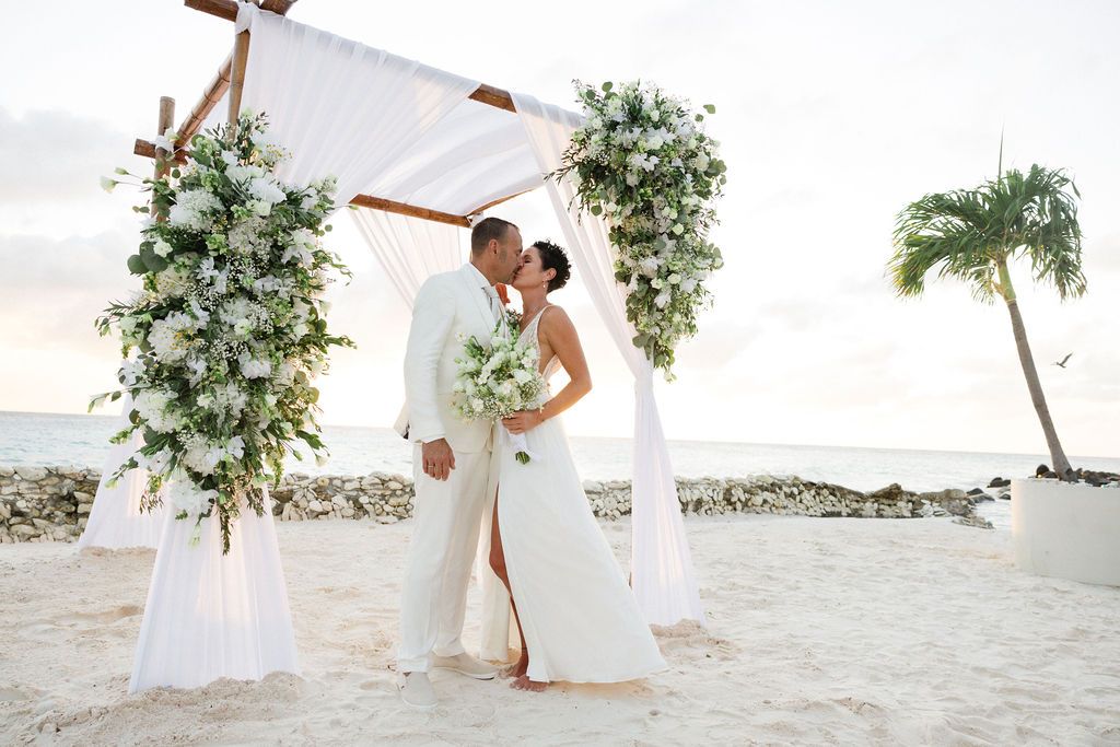 Newlyweds share first kiss under elegant white draped arch adorned with tropical greenery on beach.