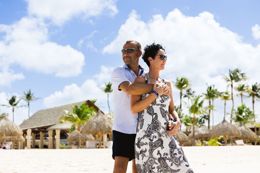 A couple embraces on a tropical beach with palm trees and thatched huts behind them.