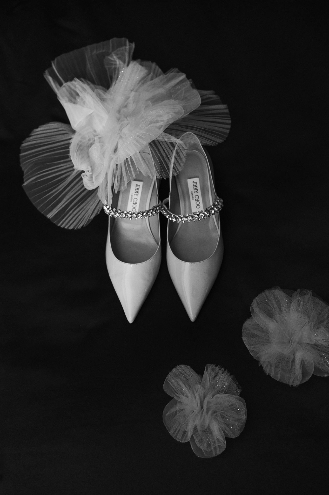 Black and white photo of elegant Jimmy Choo bridal heels with crystal straps beside delicate tulle flowers.