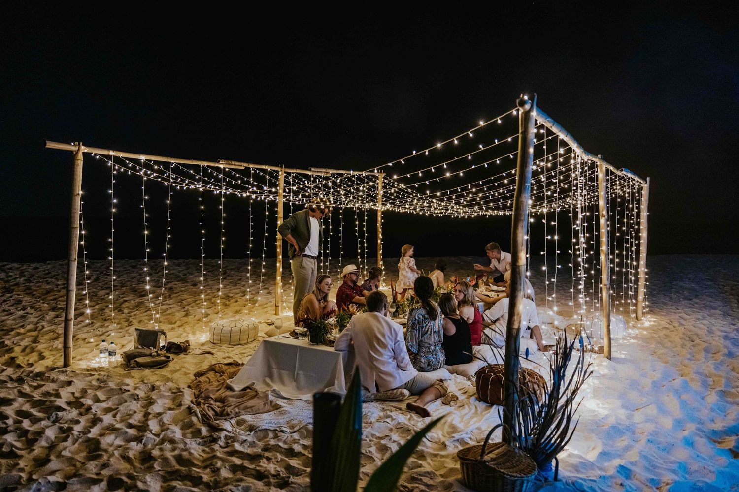 Group dining under fairy light canopy on the beach at night during outdoor celebration.