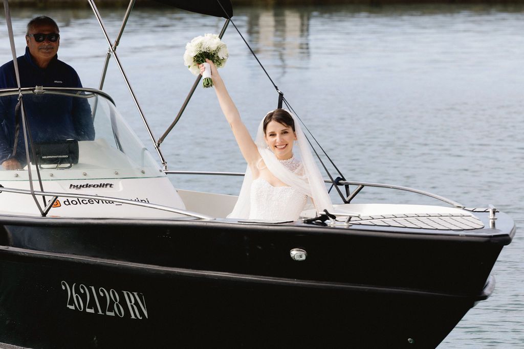 Joyful bride in white gown raising bouquet while arriving by motorboat on scenic waterway.