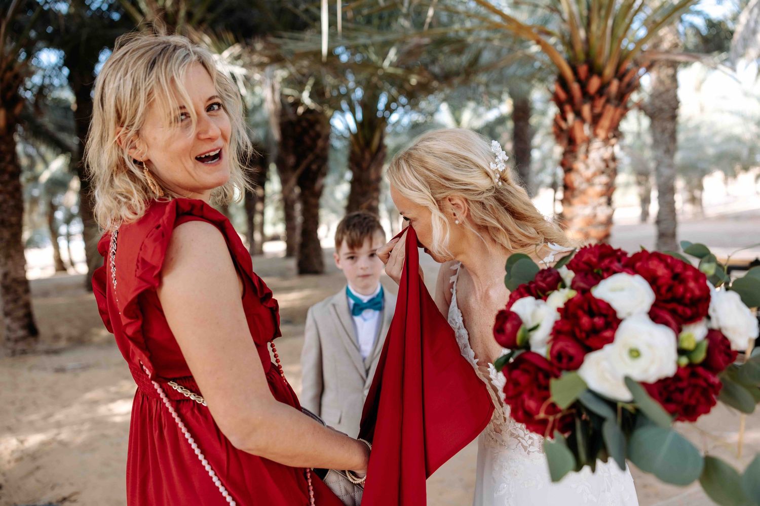 Blonde woman in red dress laughing with bride holding red and white bouquet among palm trees.
