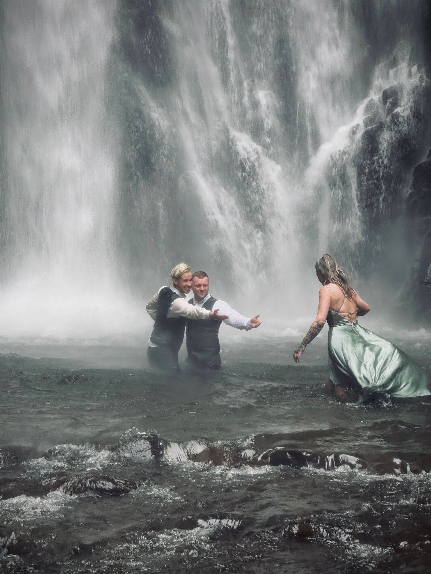 Wedding party wading through a misty waterfall pool, bride in sage green gown reaching toward grooms.