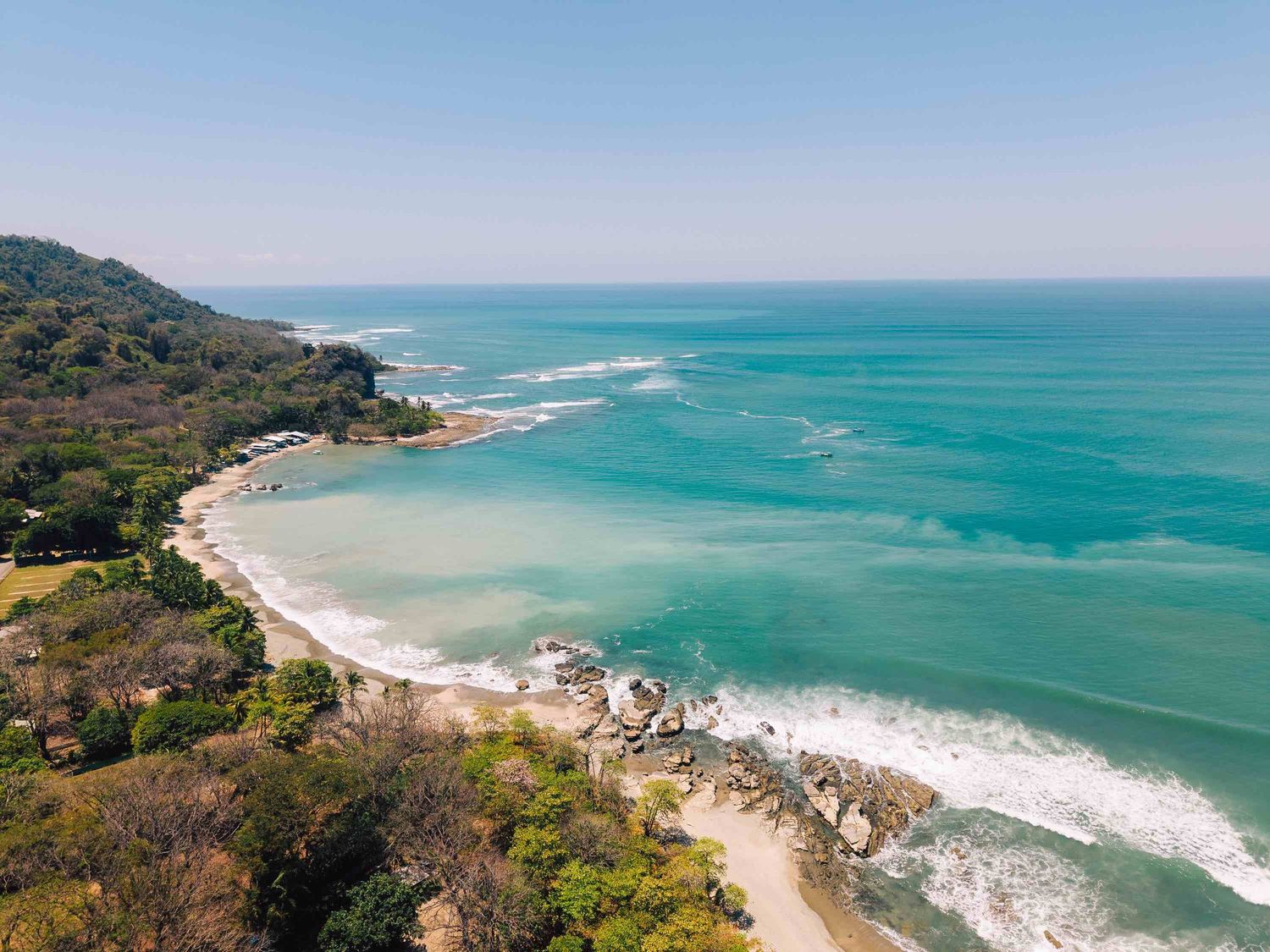 Aerial view of a tropical beach with turquoise waters, lush green trees, and rocky coastline under a clear blue sky.