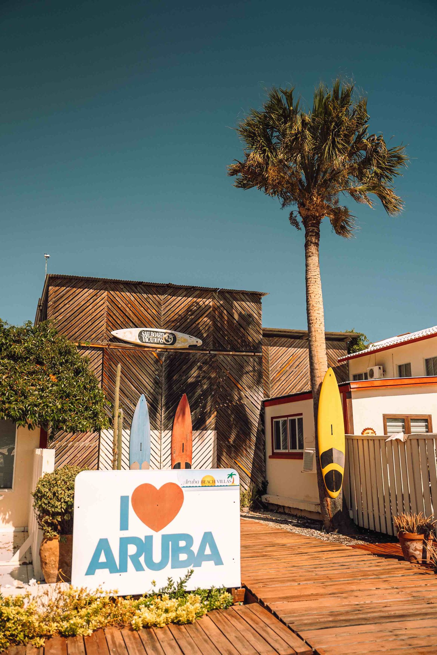 I Love Aruba sign with colorful surfboards and a palm tree outside a wooden beach shop in Aruba.