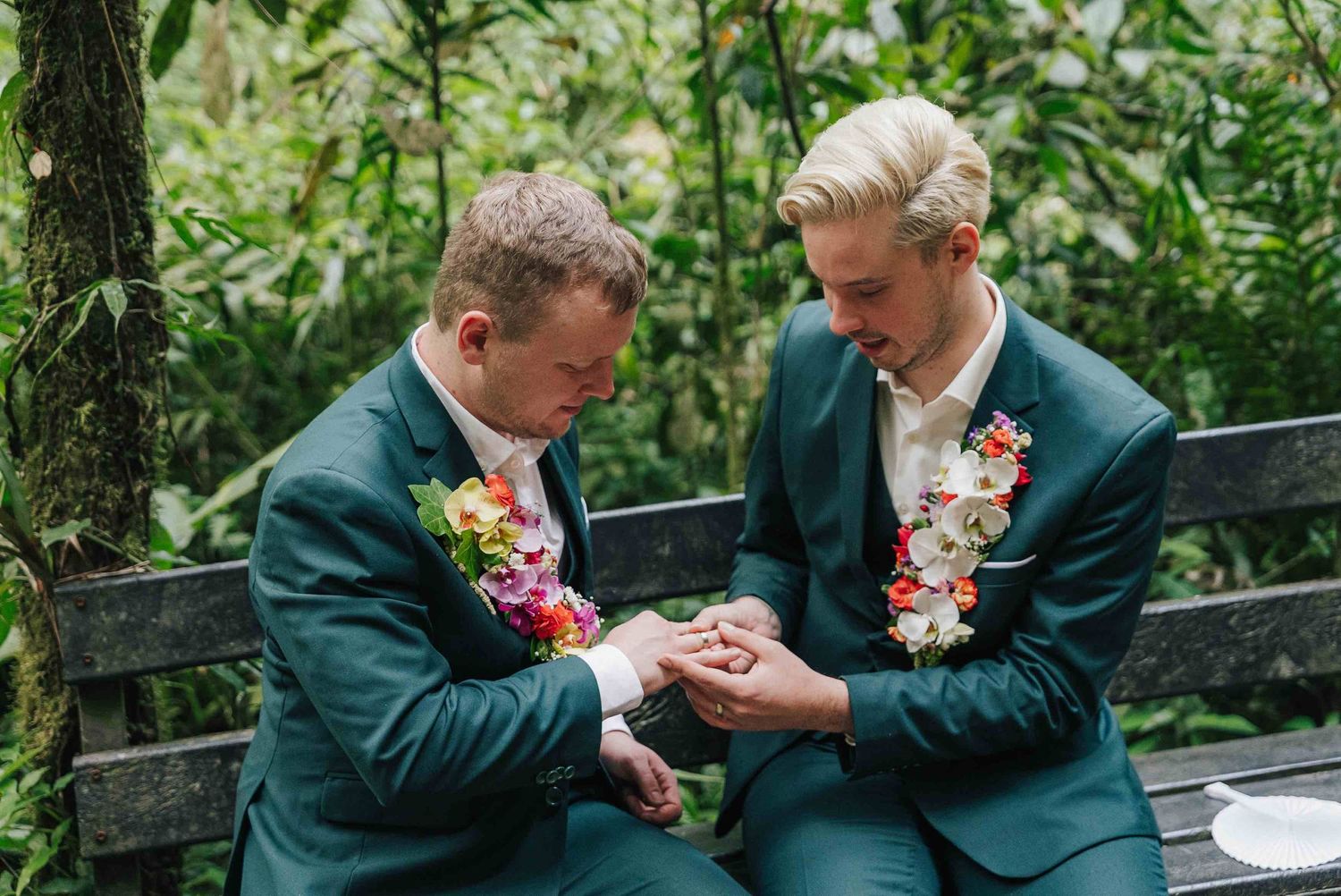 Two grooms in teal suits with floral boutonnieres exchange rings on a bench surrounded by lush greenery.