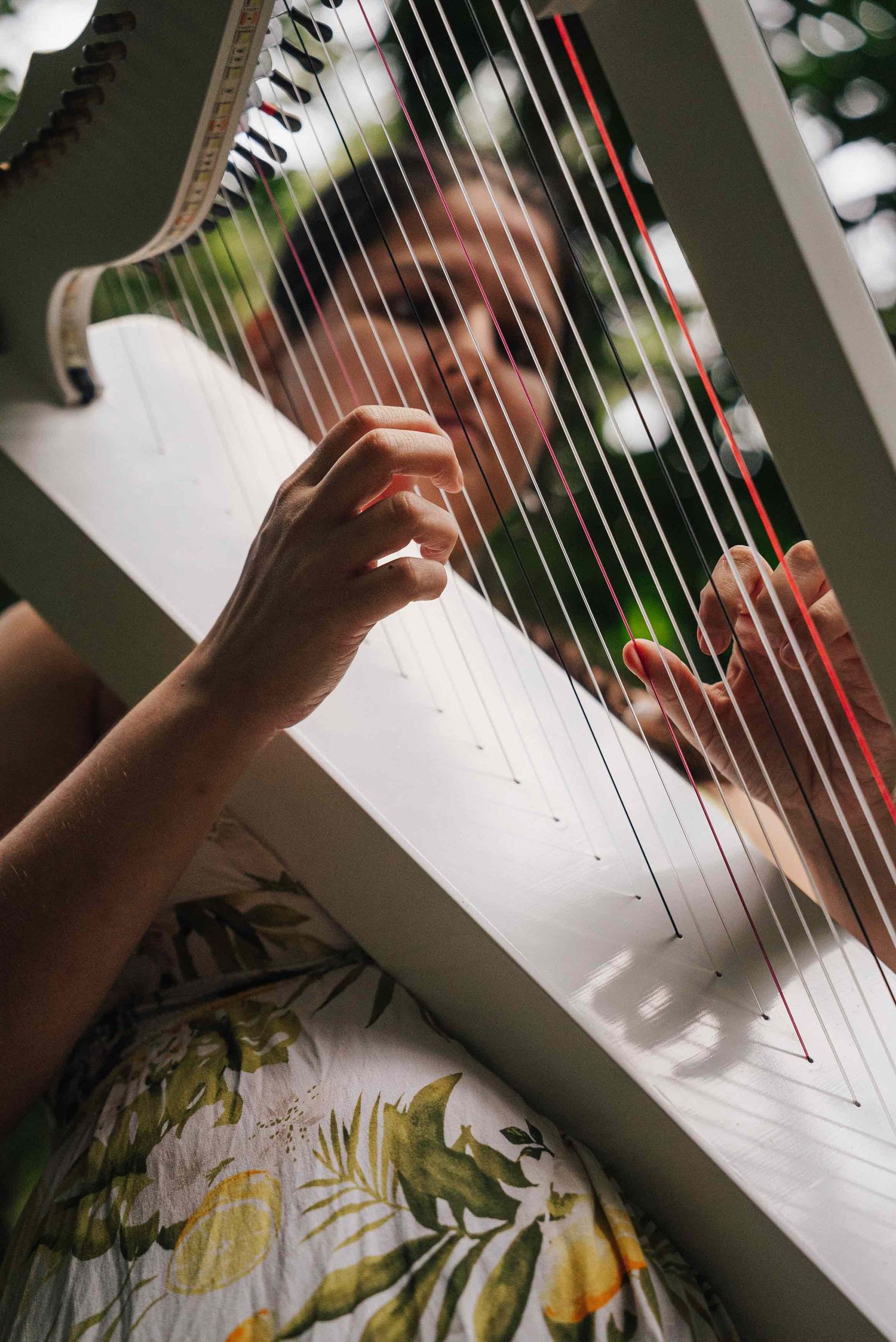 Woman playing a white harp outdoors, fingers plucking strings, wearing a floral dress in natural light.