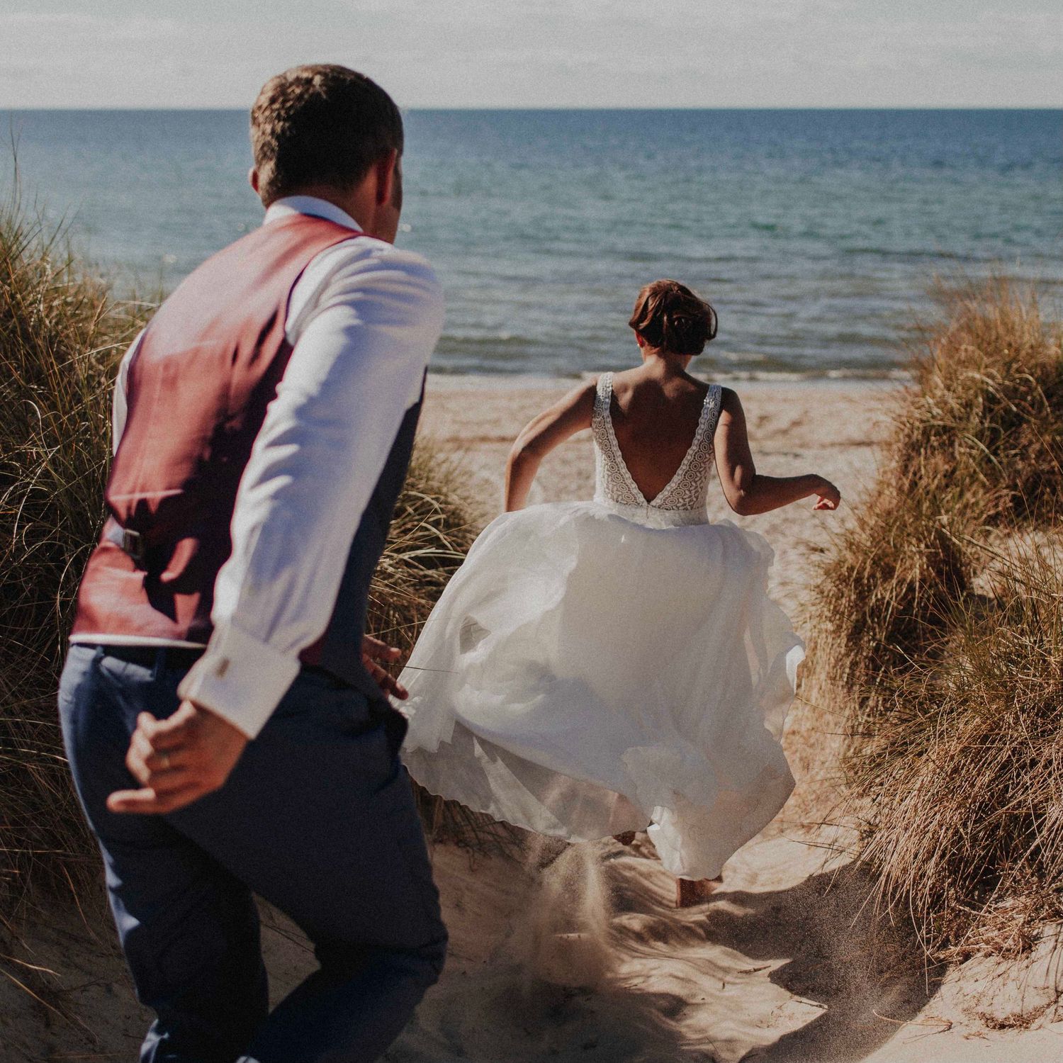 Bride and groom walking through sand dunes toward ocean beach on wedding day.