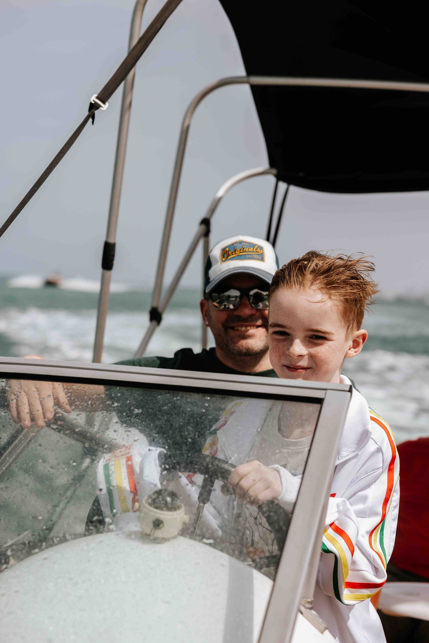 Father and young red-haired son smiling together at the helm of a speedboat on choppy water.