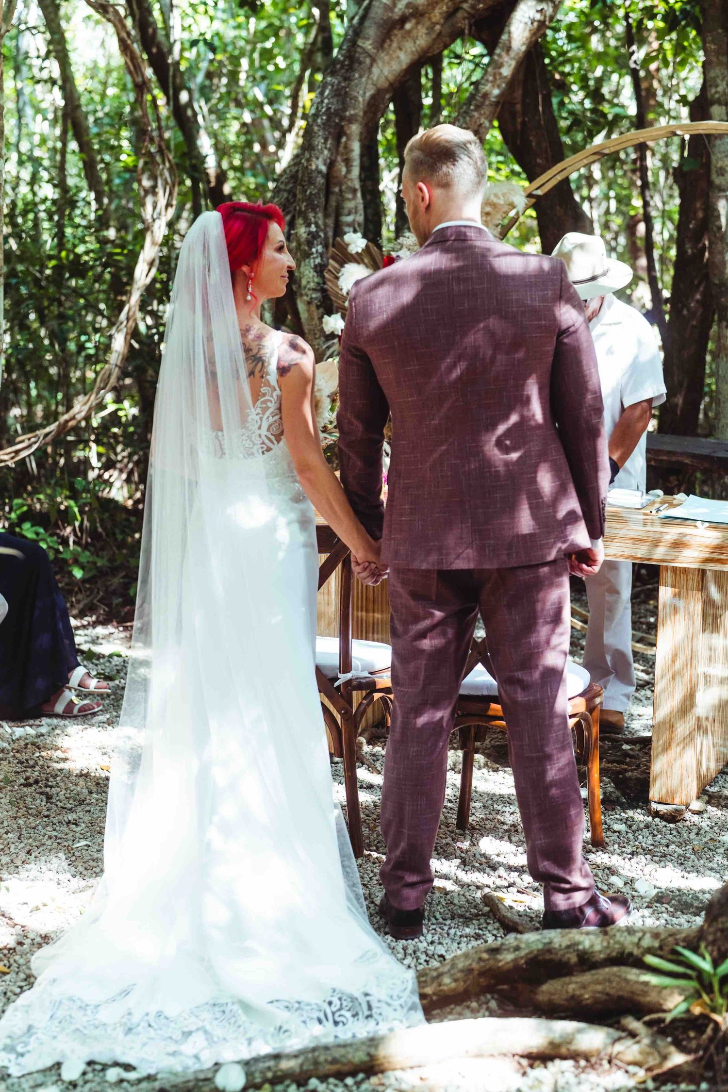 Bride in white gown and groom in purple suit holding hands at outdoor woodland wedding ceremony.