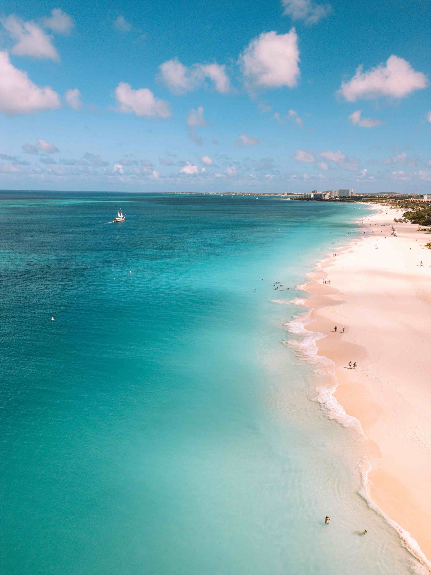 Aerial view of a stunning turquoise Caribbean beach with white sand, calm waters, and a sailboat under a blue sky.