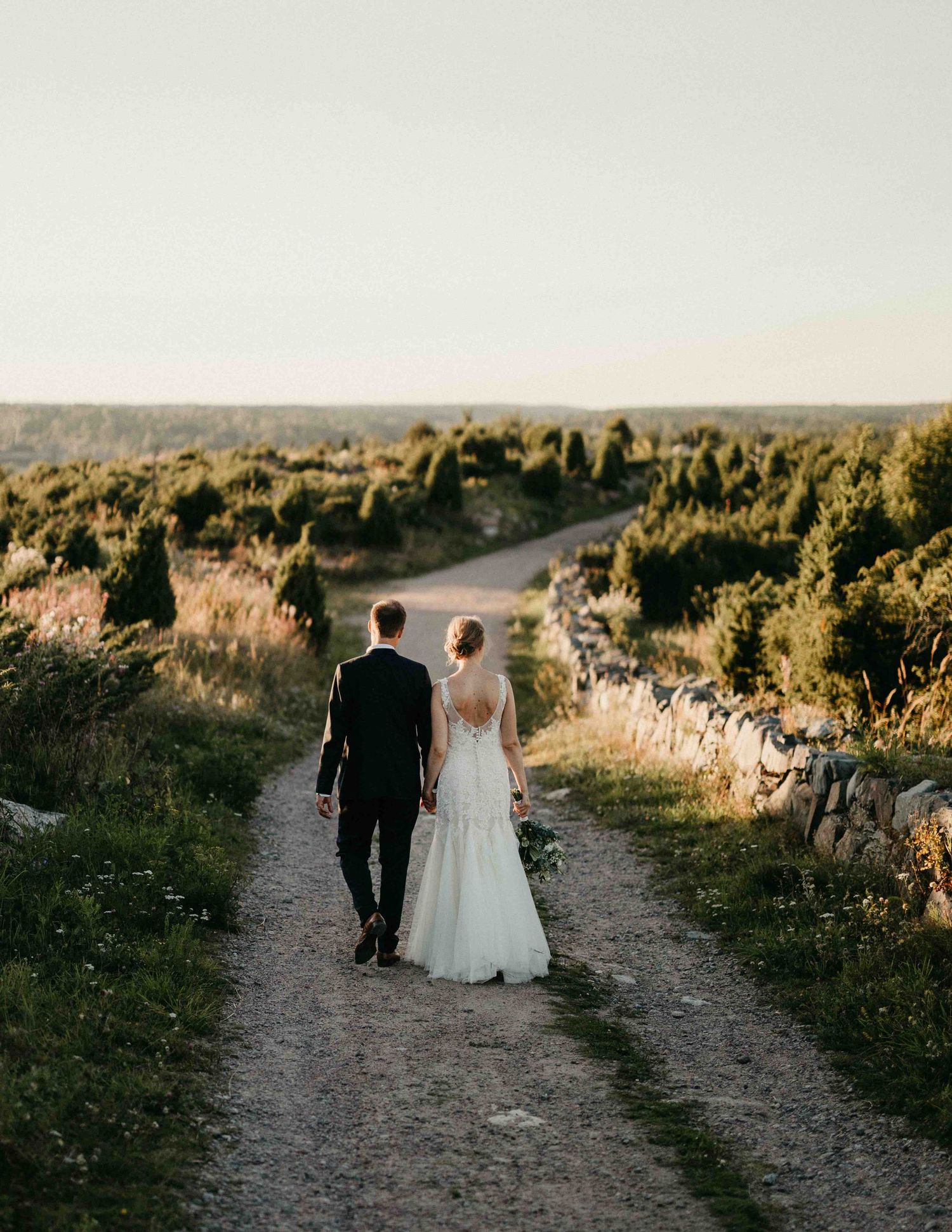 Newlywed couple walking hand in hand down a scenic countryside path at golden hour sunset.