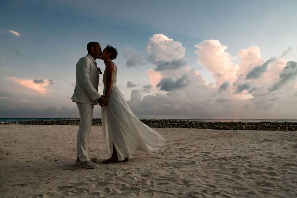 Bride and groom share a romantic beach kiss at sunset in white wedding attire with dramatic cloudy sky backdrop.