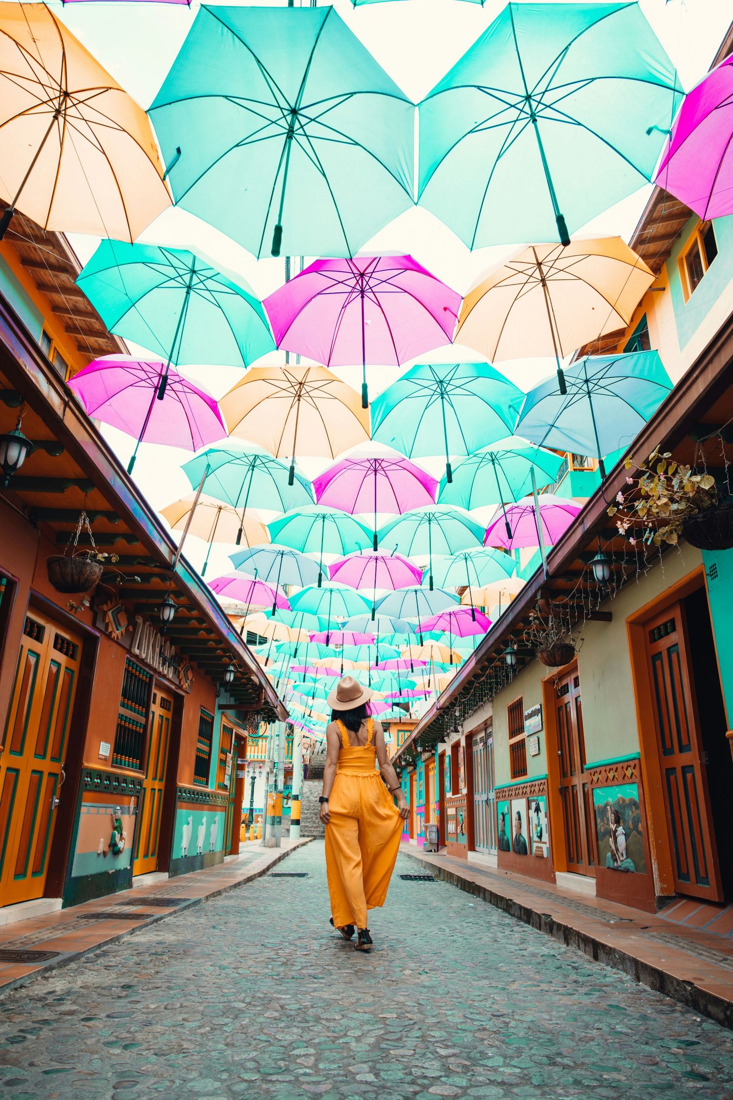 Woman in yellow jumpsuit walking under colorful umbrella canopy on charming cobblestone street.
