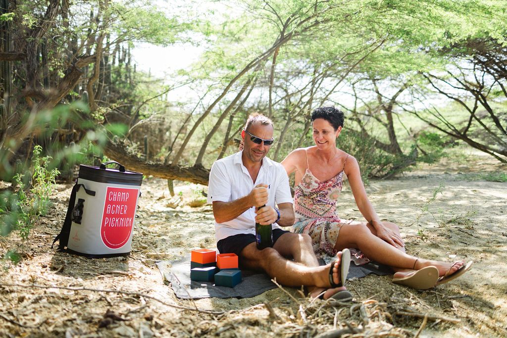 Couple enjoying drinks and a picnic under shady trees in a tropical outdoor setting.