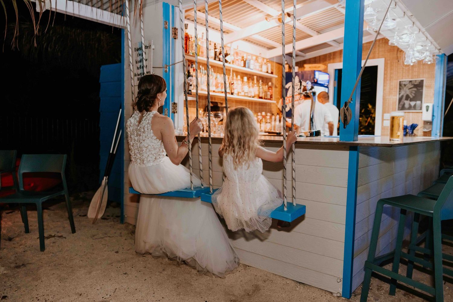 Bride and flower girl sit on swing seats at a charming beach bar with blue wooden accents.